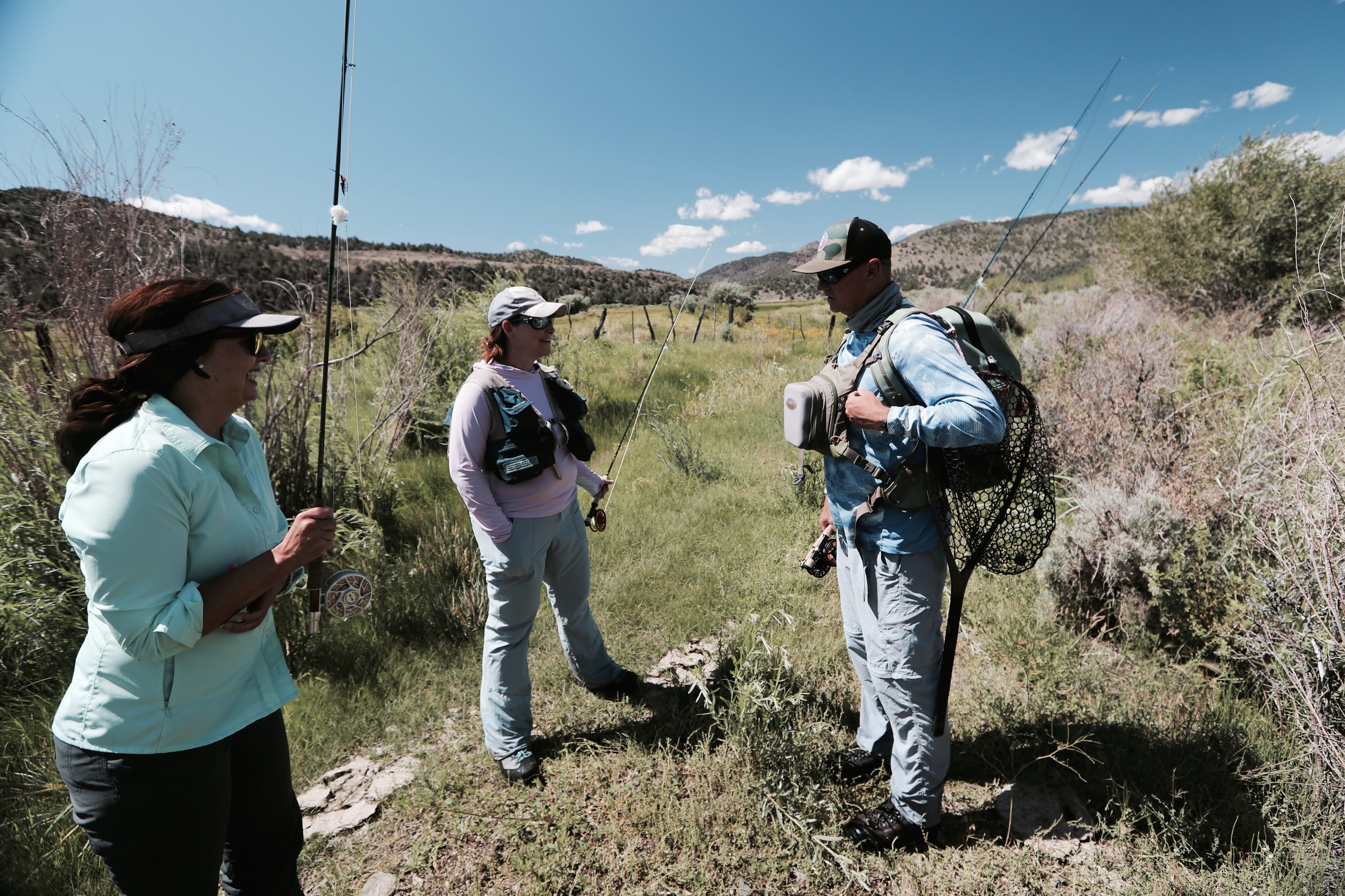 Guide instructing two women anglers on the river, net on back, sagebrush meadow behind them